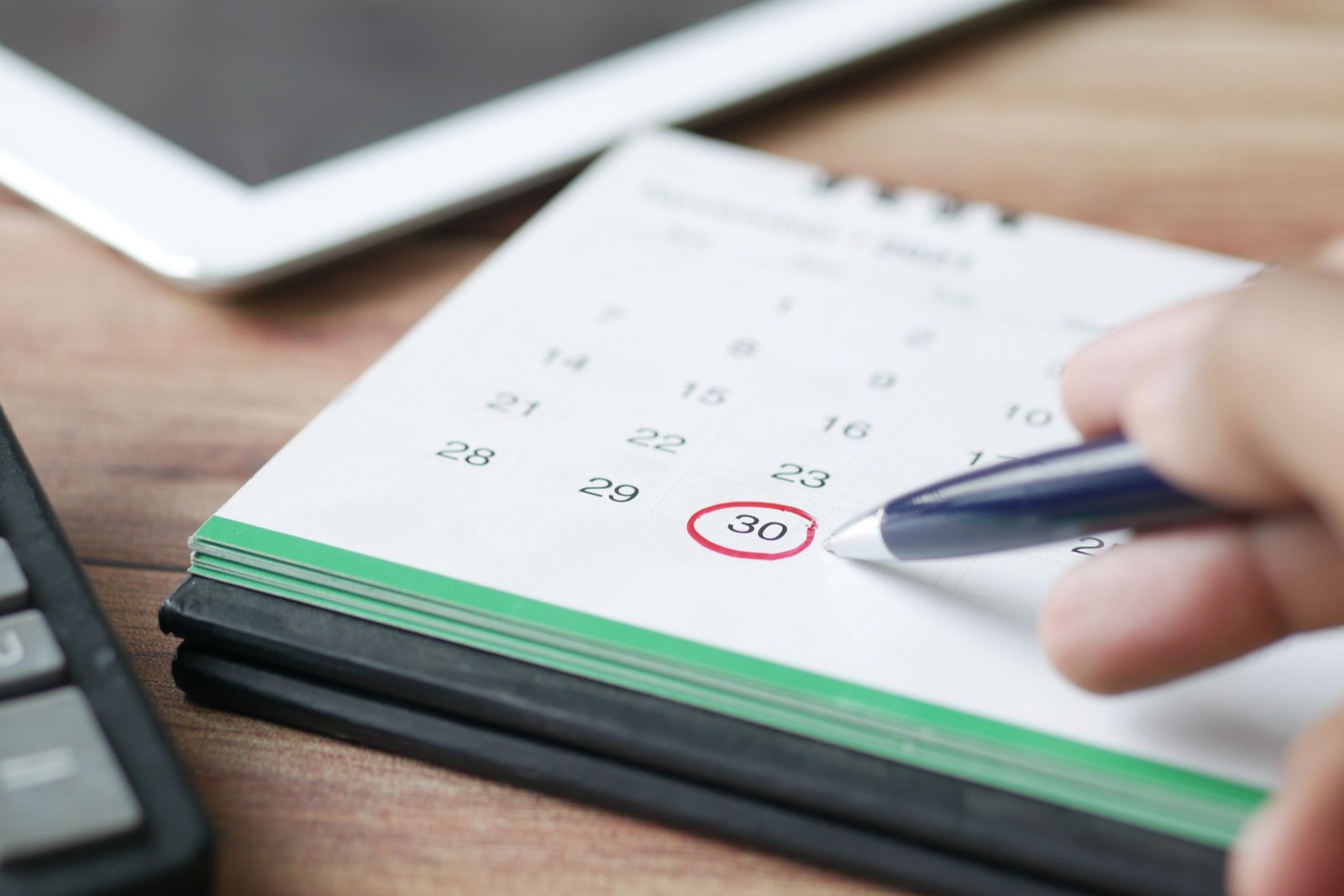 Hand with a pen pointing at a date circled in red on a desk calendar