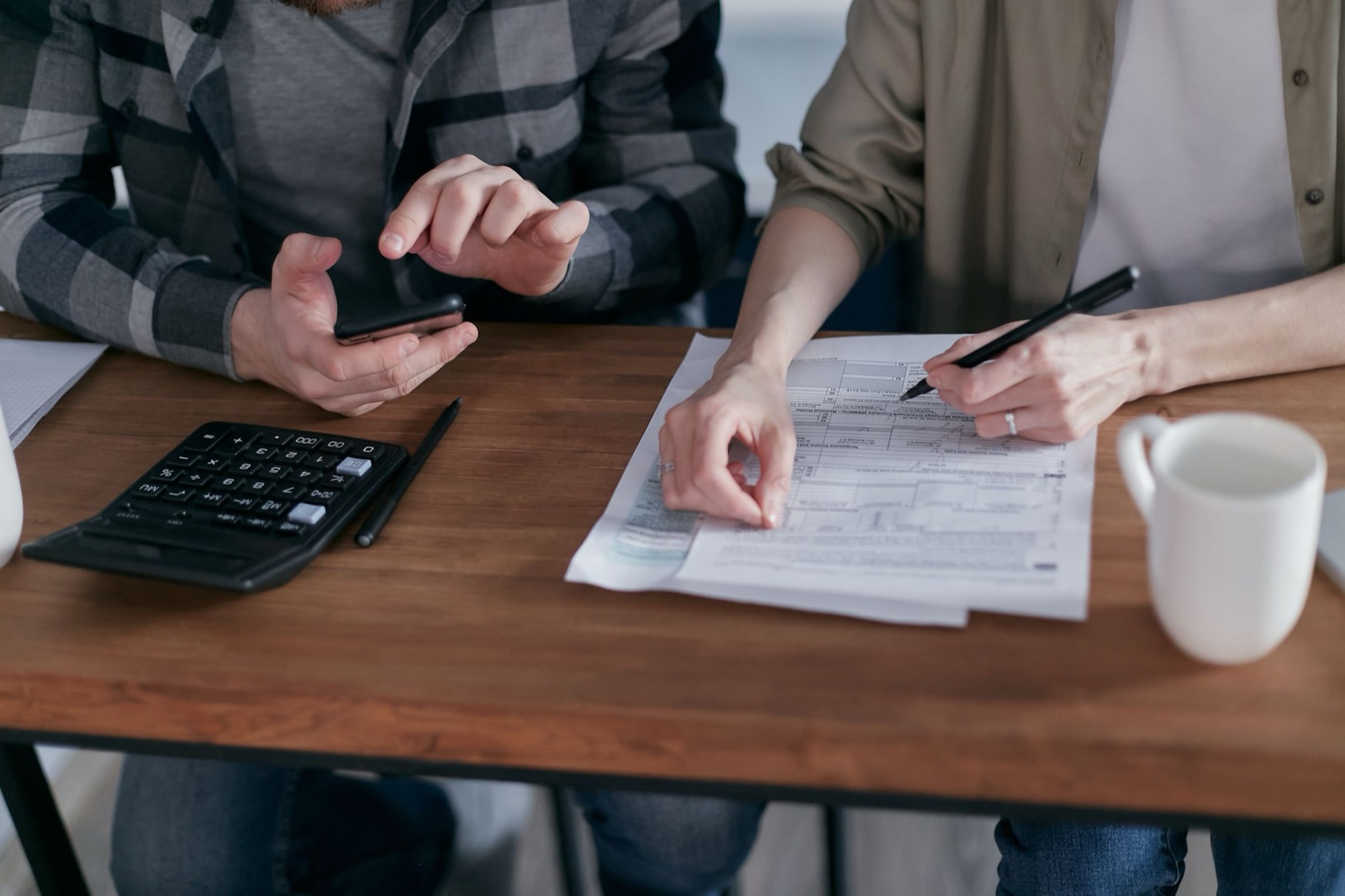 Two people at a wooden table filling out a tax form with a pen, calculator, phone and coffee mug