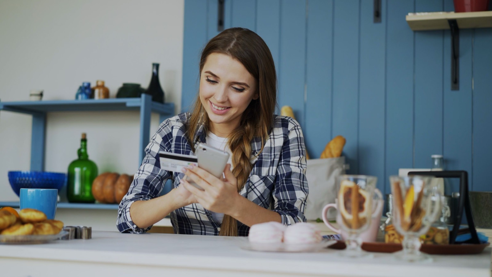 Smiling woman in a home kitchen holding a payment card and paying on her phone