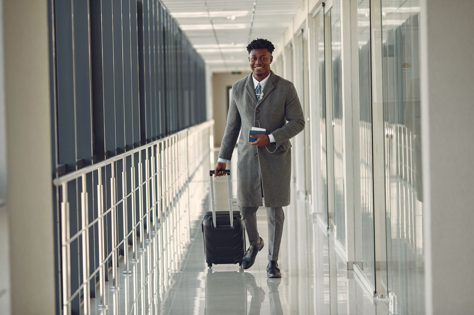 Smiling man in a gray coat walking through an airport corridor with a suitcase and passport