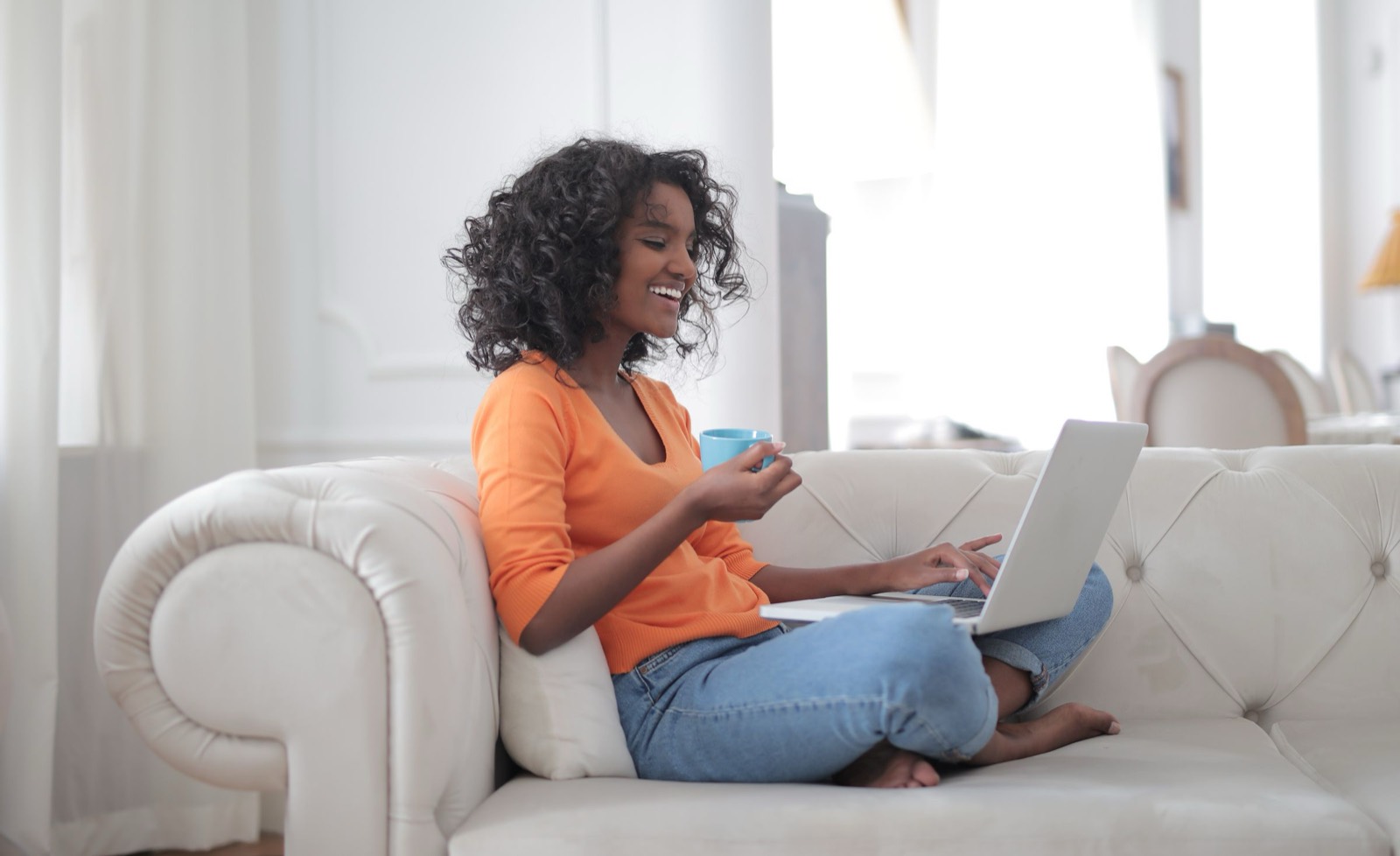 Smiling woman sitting on a white couch with a laptop and a cup of coffee in a bright room