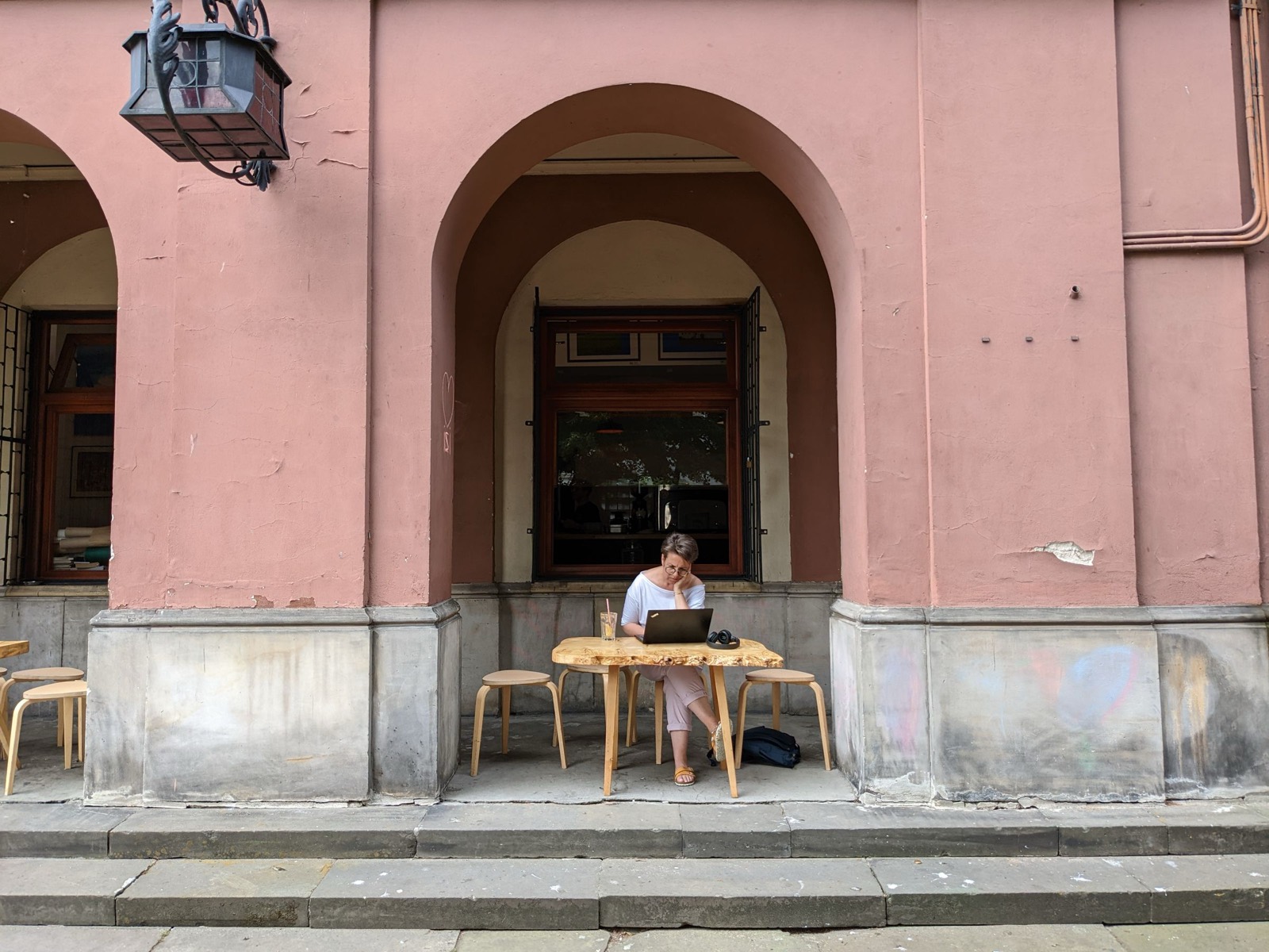 Woman working on a laptop at an outdoor cafe table under pink arches in a European city